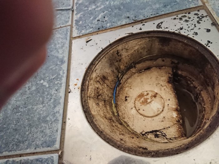 Öppen golvbrunn omgiven av blå kakelplattor i ett badrum som behöver renovering, synlig klämring ca 4cm upp.