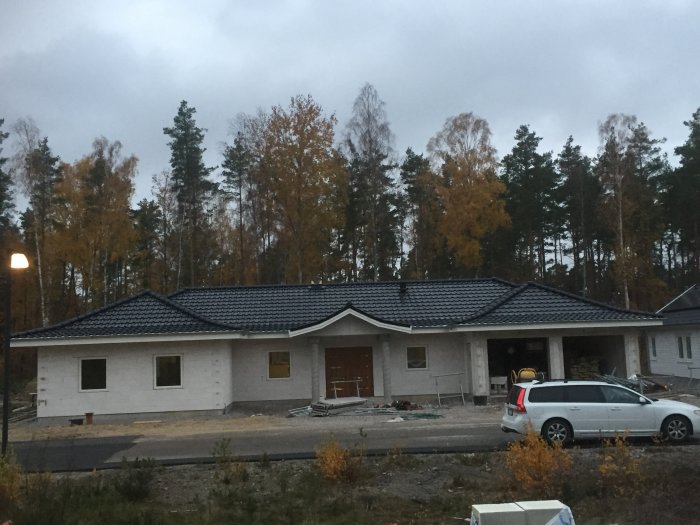 En nybyggd enplansvilla med tegeltak och vit fasad under skymningen, framför skogsmiljö.