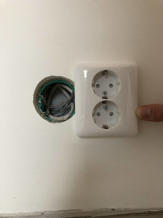 Vägg med öppen dosa bredvid monterat dubbeluttag och ett finger som pekar på installationen.