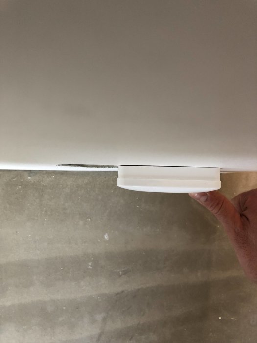 En hand håller en gipsväggskant intill en ojämn golvsockel i ett ofärdigt sovrum, markerande en konstruktionsdetalj.