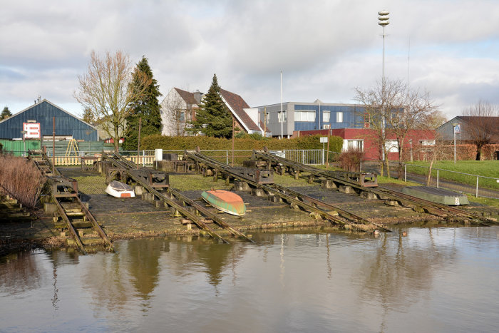 Övergivet skeppsvarv med gamla båtar och förfallna ramper vid vattnet, omgivet av byggnader och grönska.