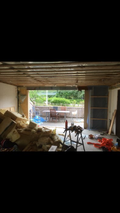 Renoveringsprojekt med synliga takstolar och isolering, verktyg och byggmaterial på golvet, öppet skjutparti ut mot terrass.