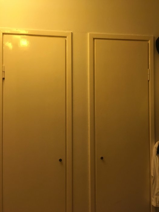 Två inbyggda garderober i hall med slutna dörrar, gul väggfärg med synliga reglar ovanför.