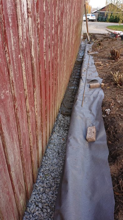 Grundarbete längs en byggnad med markduk och makadam före betongblandning.