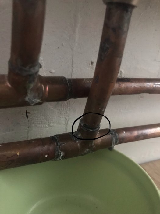 Inringad skarv på ett kopparrör som läcker vatten nära en vägg, med droppfat nedanför.