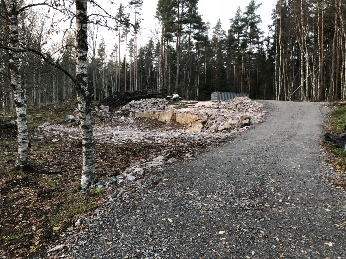 Grusväg som leder till en uppgrävd tomtyta med högar av sprängsten och natur omkring.