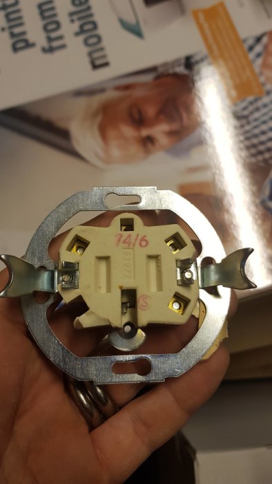 Hand håller en gammal elektrisk brytare med synliga anslutningsterminaler märkta med bokstäver och siffror.