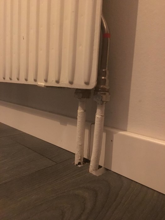 Vit radiator ansluten med ett par misstänkta enrörssystemsrör mot golv och vägg.