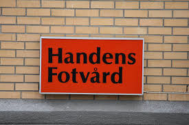 Röd skylt med texten "Handens Fotvård" mot en gul tegelvägg.