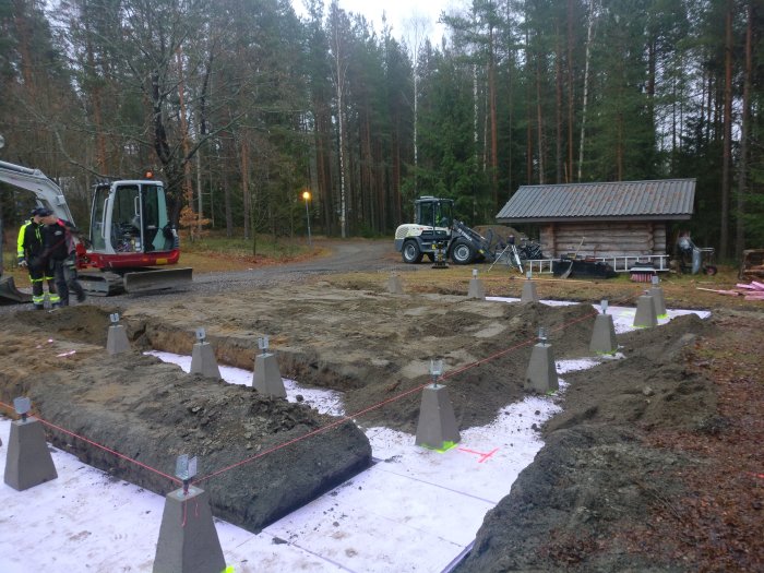 Byggarbetsplats med grovfyllt område mellan betongplintar, grävmaskin, lastare och skog i bakgrunden.