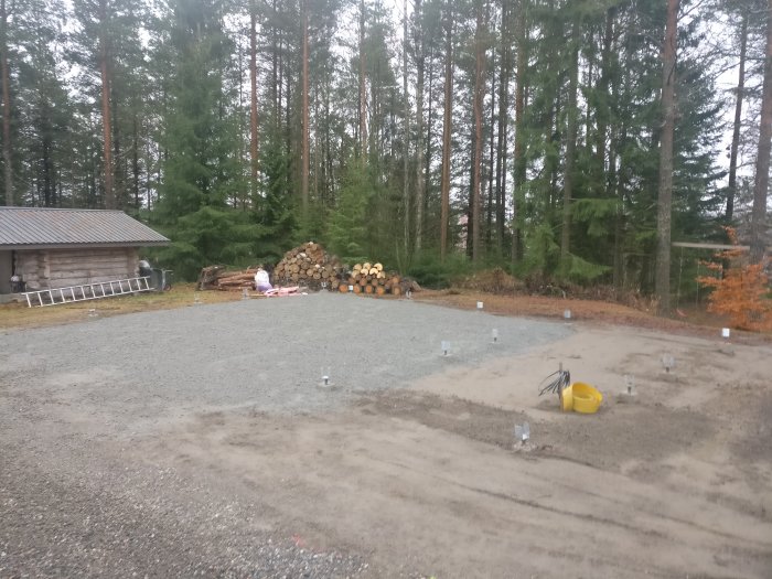 Nybyggd husgrund med markarbeten klara och fundamentpelare på en grusad tomt med skog i bakgrunden.