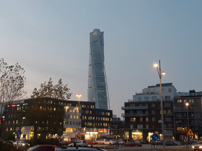 Skymningsvy över stad med ett framträdande torn i bakgrunden och byggnader och parkerade bilar i förgrunden.