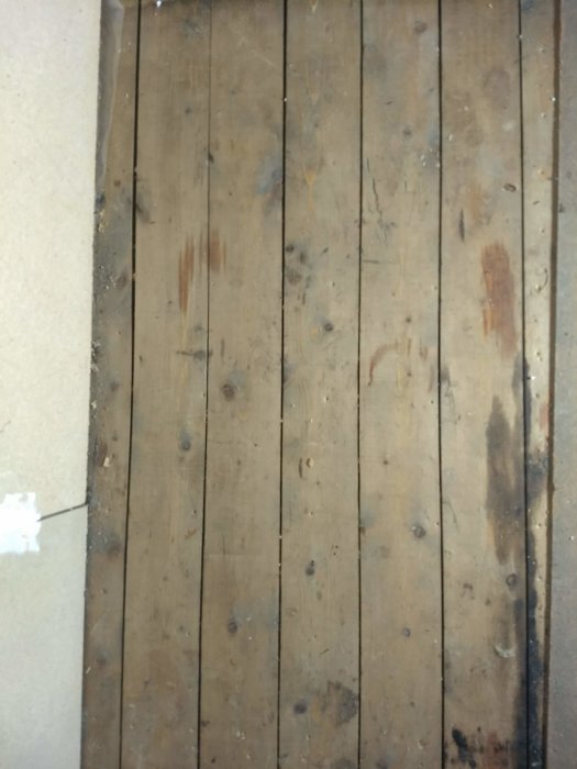 Gammalt trägolv i behov av renovering med synliga nötningar och fläckar, intill en vit väggkant.