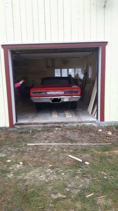 Klassisk röd bil backas ut från garage på träramper, person i bakgrunden guidar.