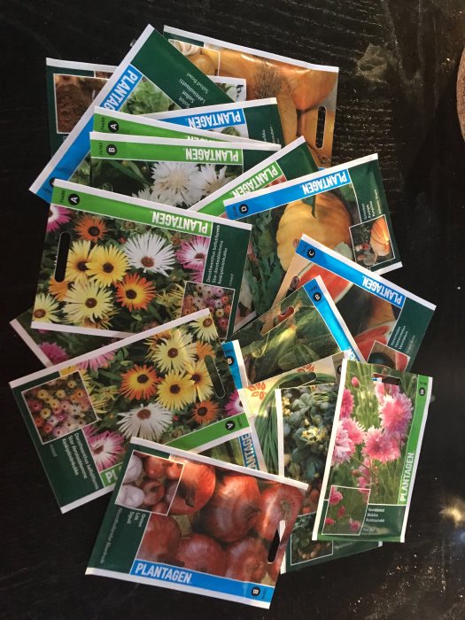 Hög med olika fröpåsar från Plantagen med bilder på blommor och grönsaker på en mörk bakgrund.