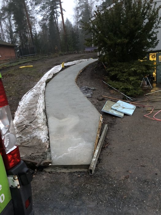 Nybyggd slingrande betonggång med formar i en trädgård, arbetsmaterial synligt, skymningsljus.