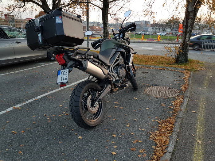 Nyare motorcykel parkerad med stor förvaringsbox på baksidan, vid vägkant omgiven av höstlöv.