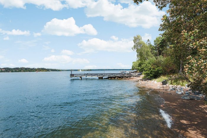 Tranquil lakeside view with a boat dock, sandy shore and clear water surrounded by greenery, reflecting a scenic nature setting.