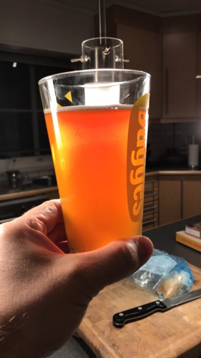 Person som håller ett glas med orange vätska, troligen hemmagjord öl, i köket.