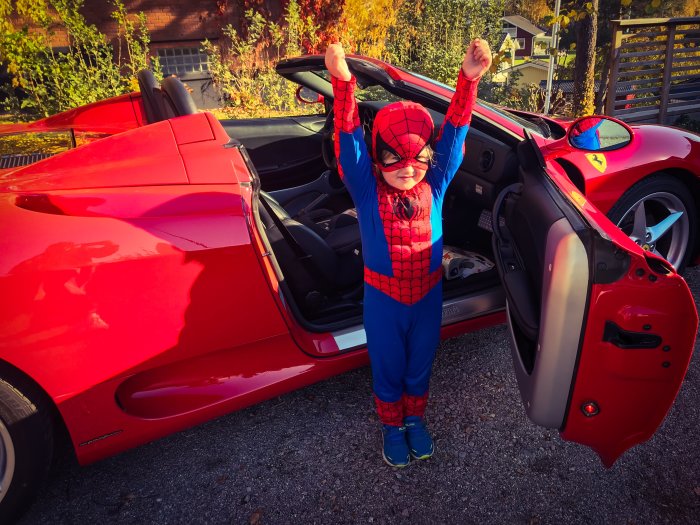 Barn utklädd till Spiderman står bredvid en röd Ferrari med öppen dörr.