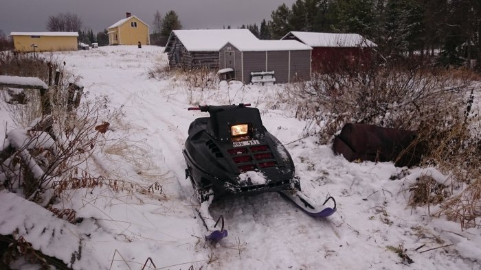 Snöskoter av märket Polaris parkerad på snöklädd mark med hus och skjul i bakgrunden.