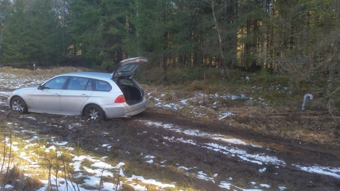 En vit BMW 325d parkerad på en lerig skogsväg med öppen baklucka.