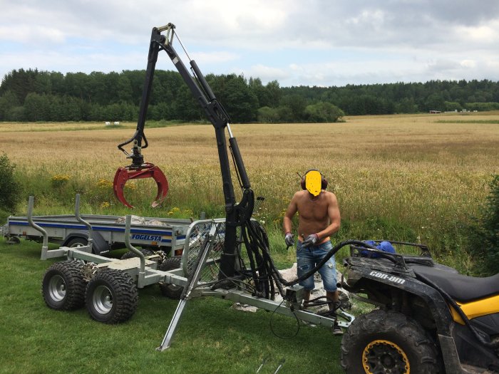 Hydraulvagn med kranlyft monterad på släpvagn och person som arbetar med fyrhjuling på äng.