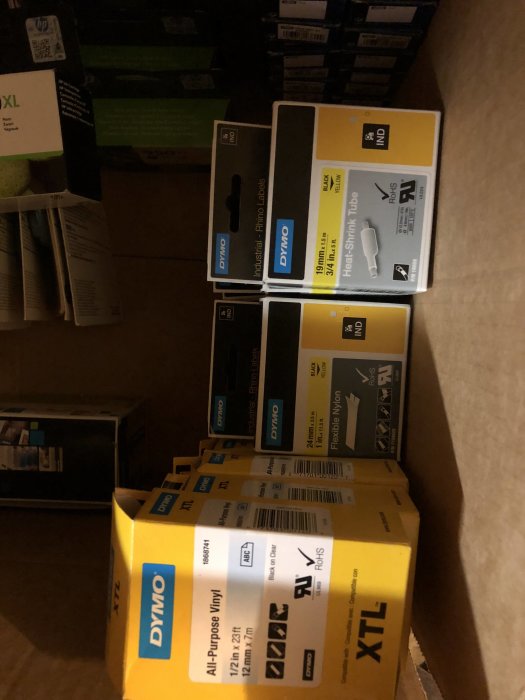 Flera förpackningar av Dymo etikettrullar och krympslang staplade i en kartong.