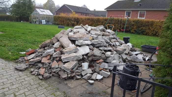 Hög med blandat byggavfall på en trädgårdsgång, inklusive betong och tegel, med växthus i bakgrunden.