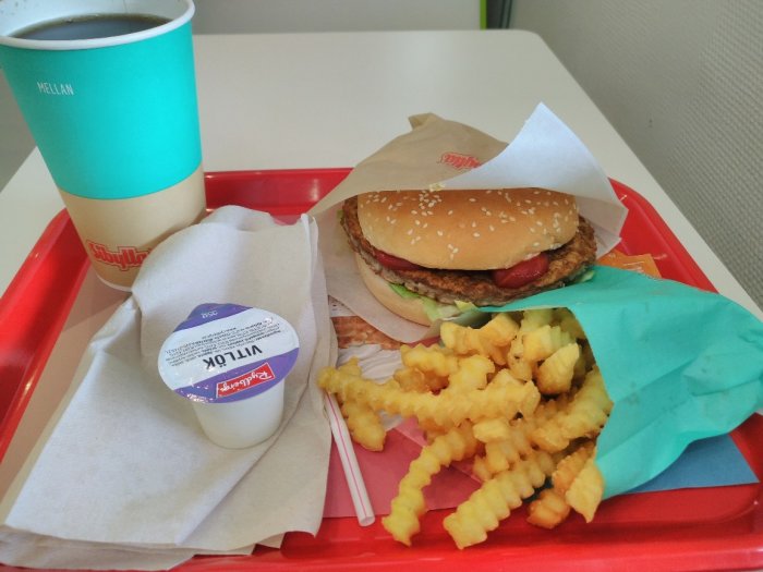 Hamburgare med bröd, grönsaker och köttpuck på pappersförpackning, krinkle cut pommes frites, mjölk och yoghurt på röd bricka.