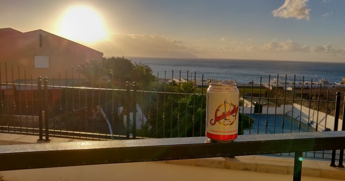 Solnedgångsutsikt över havet med ölburk på balkongräcket, pool och byggnader i förgrunden.