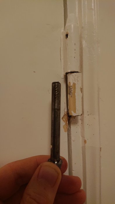 Hand håller en gammal rostig dörrgångjärnspinne bredvid en vitmålad dörrkarm.