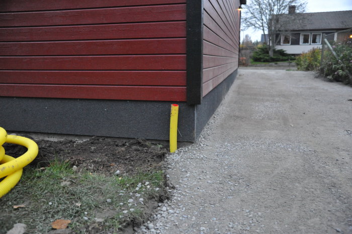 Gult elkabelskyddsrör sticker upp vid rödmålad trävägg och betongplatta med grusväg intill.