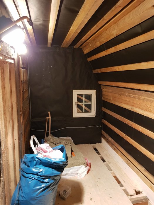 Oisolerad inomhusmiljö under renovering med synliga träreglar på vägg och snedtak, och isoleringsmaterial i säck.