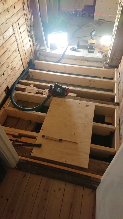 Pågående renovering av ett badrumsgolv med öppet bjälklag och framlagda plywoodskivor, verktyg och arbetsbelysning.