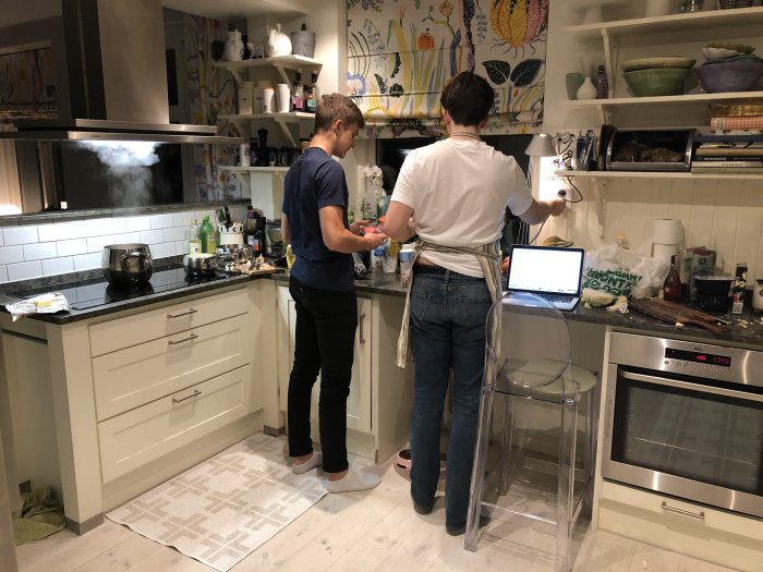 Två personer lagar mat i ett rörigt kök med moderna vitvaror och färgglad tapet.