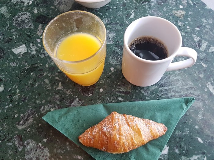 Frukost bestående av en croissant på en grön servett, ett glas apelsinjuice och en kopp kaffe på marmorbord.