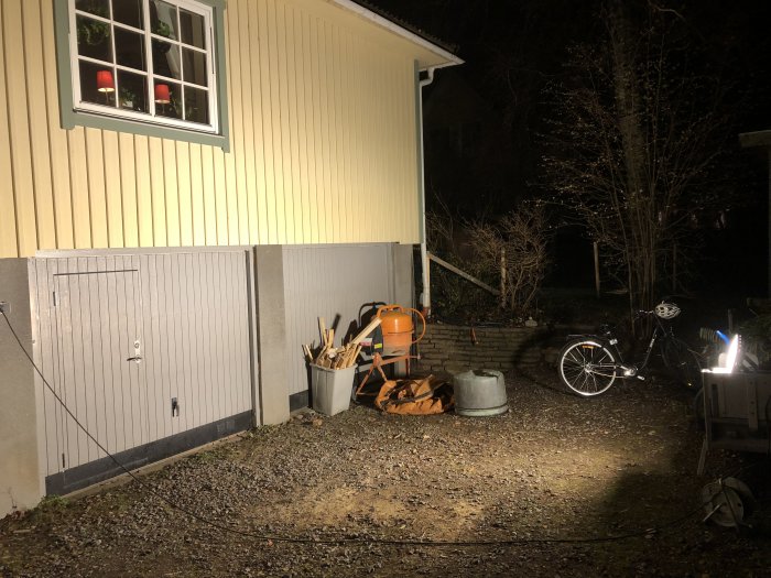 Uppvärmt garage med öppen dörr och diverse verktyg, en cementblandare och en cykel upplysta av en 300w lampa på kvällen.