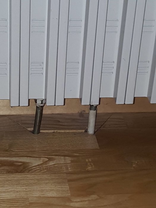 Vit radiator med två synliga rör som går in i golvet, frågeställning om rörsystem.