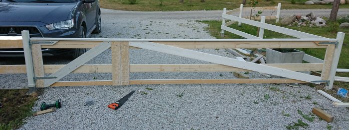 Halvfärdig vit trägrind under byggnation, med horisontella remsor och markerad stolpe med gröna fläckar, framför en grusuppfart.