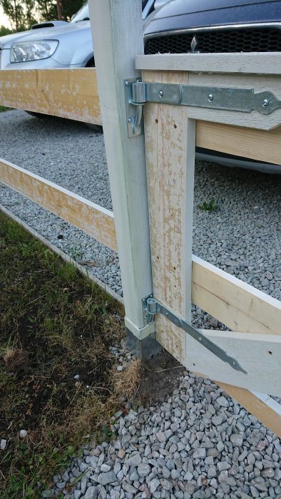 Närbild på en halvfärdig trägrind med synlig missfärgning och metallbeslag, bredvid en grusväg och en bil.