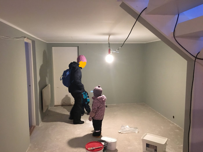 En vuxen och ett barn i rum under renovering med nyputsade väggar och oulagt golv, byggmaterial synligt.