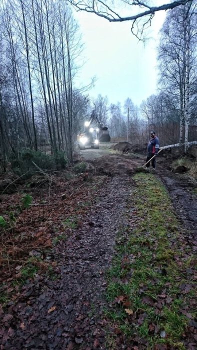 Grävmaskin i arbete med första spadtaget för ett husbygge, person i bakgrunden, skogsmiljö.