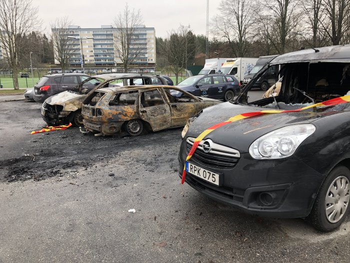 Brända bilar på en parkeringsplats med en byggnad och träd i bakgrunden.