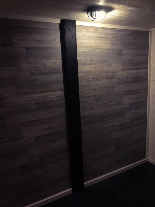 Ett nyligen färdigställt gästrum med mörkt trägolv och en vägg klädd med grå brädor, upplyst av en taklampa.