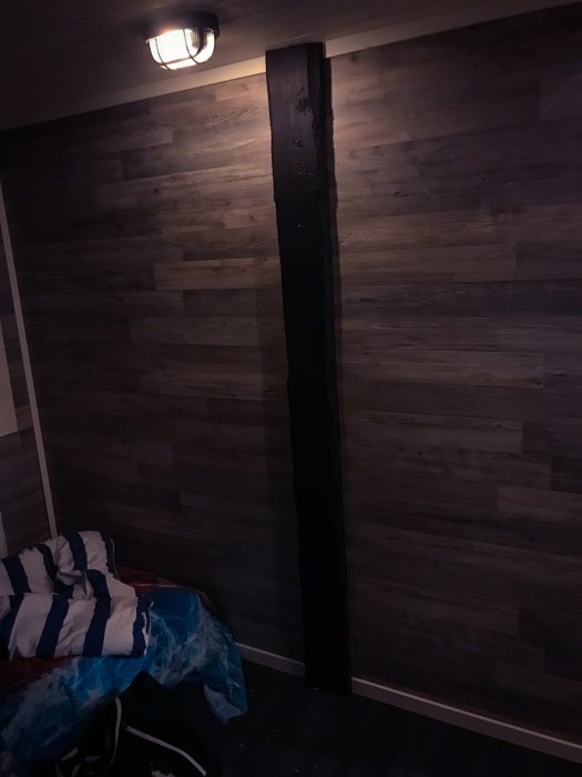 Ett hörn av ett sovrum med mörkt träliknande tapet och en tillfällig tältsäng under en vägglampa.