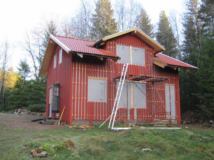 Rödmålat hus under konstruktion utan fönster och dörrar, omgivet av skog och byggmaterial.