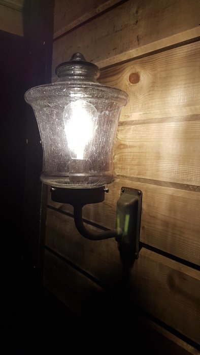 Vägglampa av oxiderad koppar med lila kupol, tänd och monterad på trävägg.