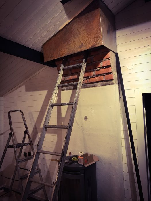 Rostig skorstenshuva som täcker en delvis murad skorsten med en stege framför i ett rum under renovering.