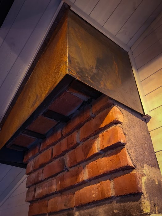 Rostskyddad skorstensdel i metall ovanpå en tegelstenspelare inomhus, med en vit trävägg i bakgrunden.